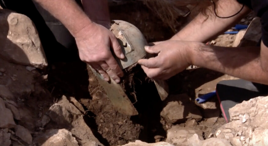 men lift a dirty gladiator-style helmet from a hole